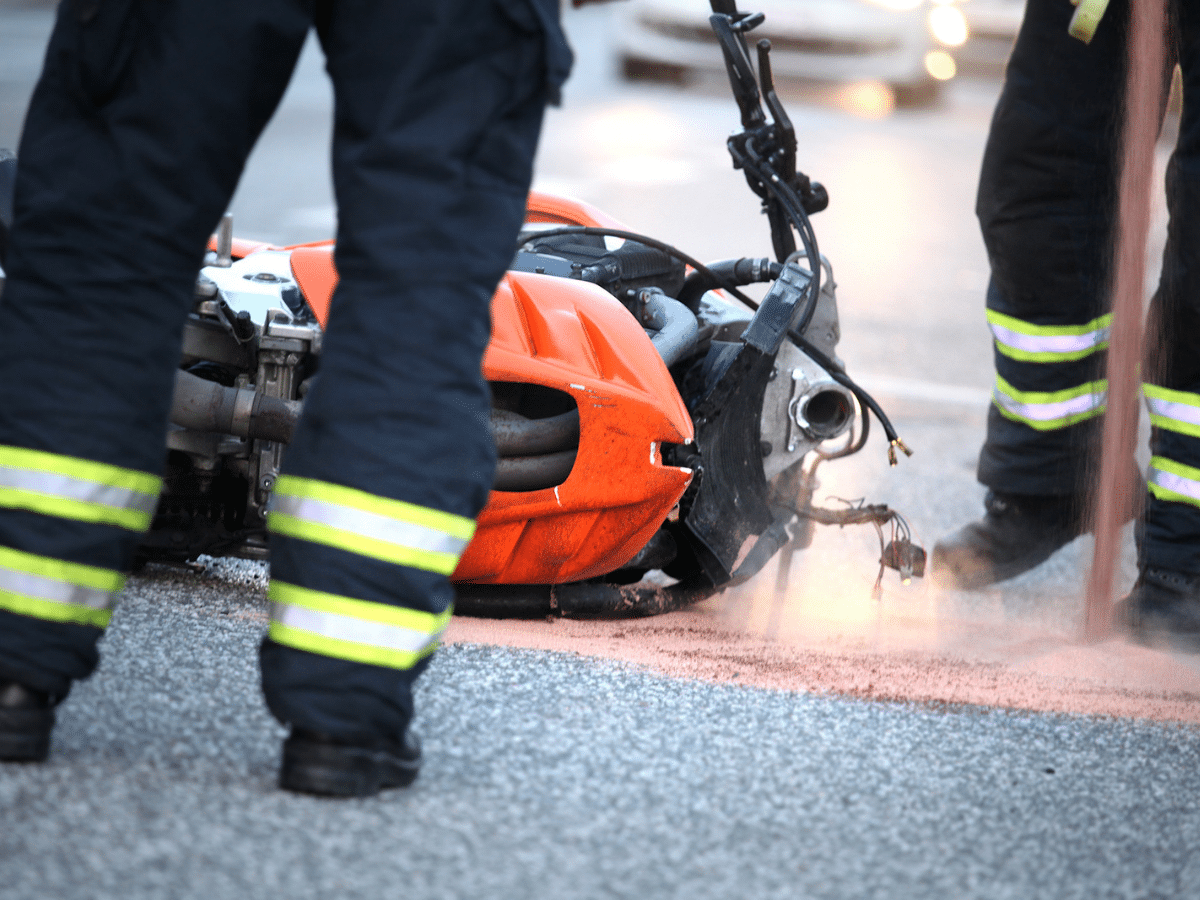 Emergency responders in high-visibility gear stand near a crashed motorcycle lying on its side in the roadway, with debris and absorbent material spread across the pavement. The image depicts the immediate aftermath of a serious motorcycle accident, highlighting the severity of impact and the types of incidents that can lead to catastrophic injuries such as traumatic brain injuries, fractures, or long-term disability.